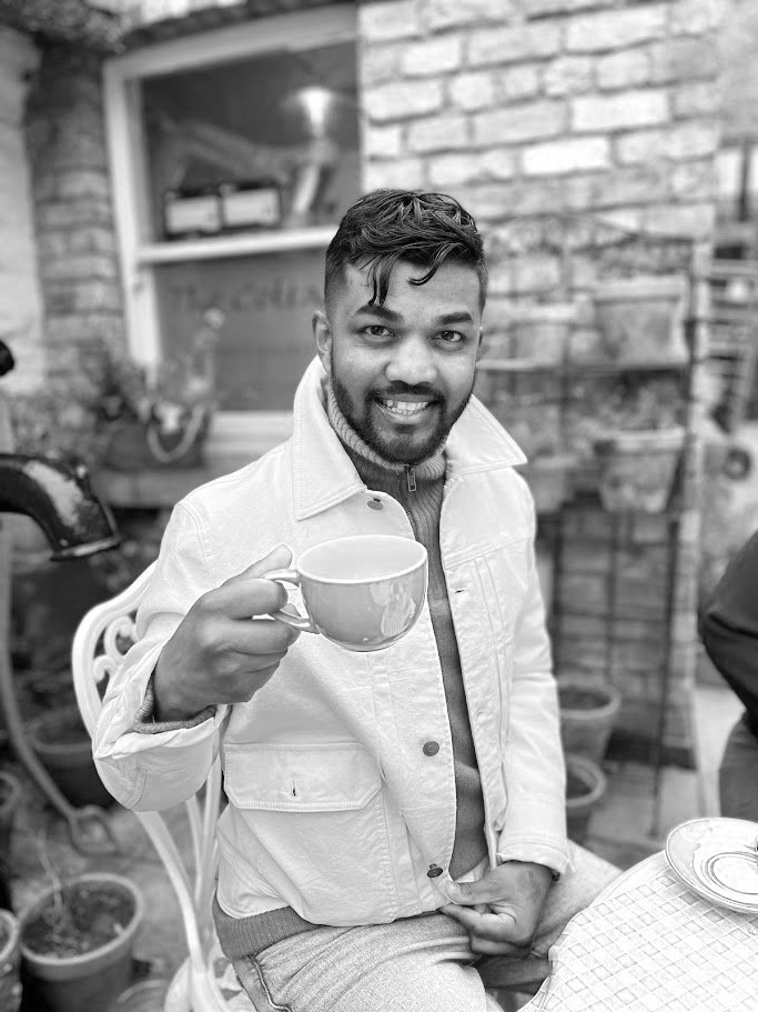 A black and white photo of John Kumar. He is smiling and holding a cup of coffee.