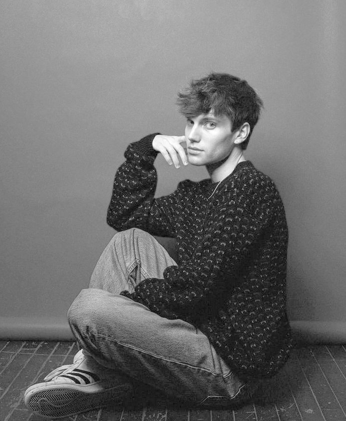 A black and white photo of Tristan Braungardt. He is sitting on the floor with his hand on his chin.