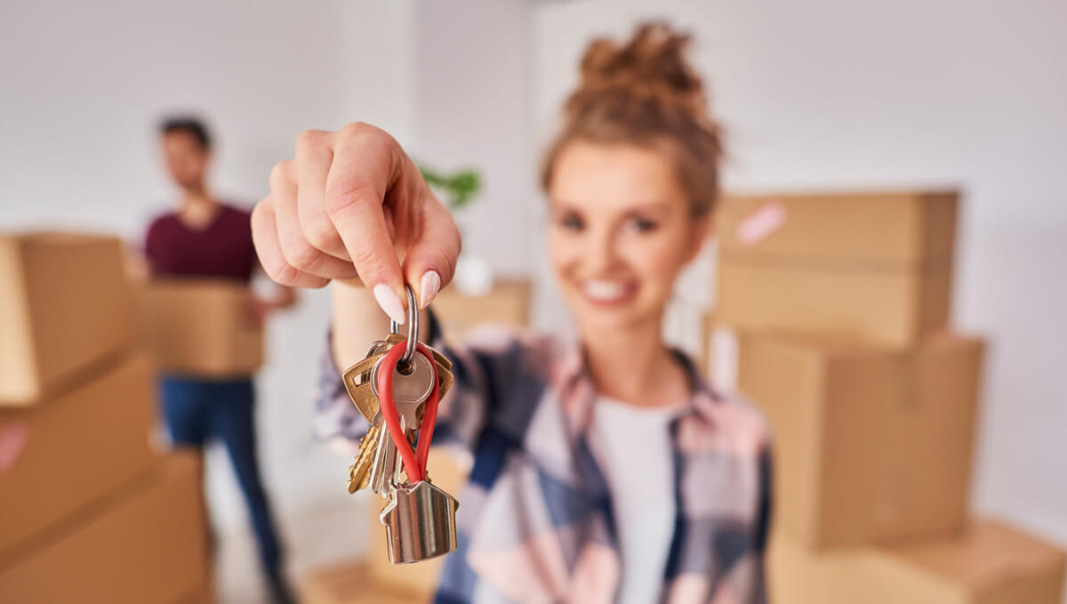 A smiling woman holds out keys to her new home. A man stands in the background, surrounded by boxes.